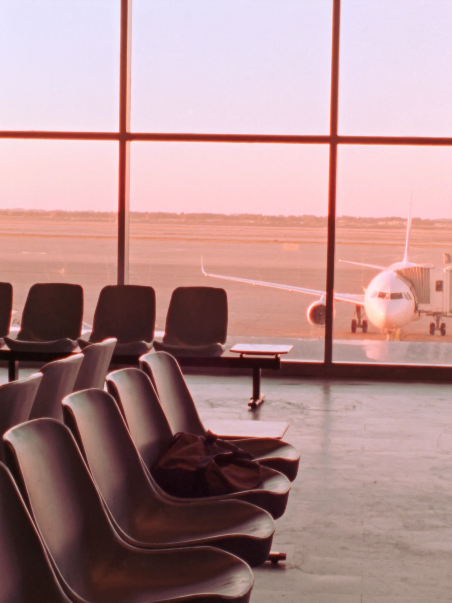 An empty airport departure gate at dawn, a bag left on a seat and a plane waiting outside