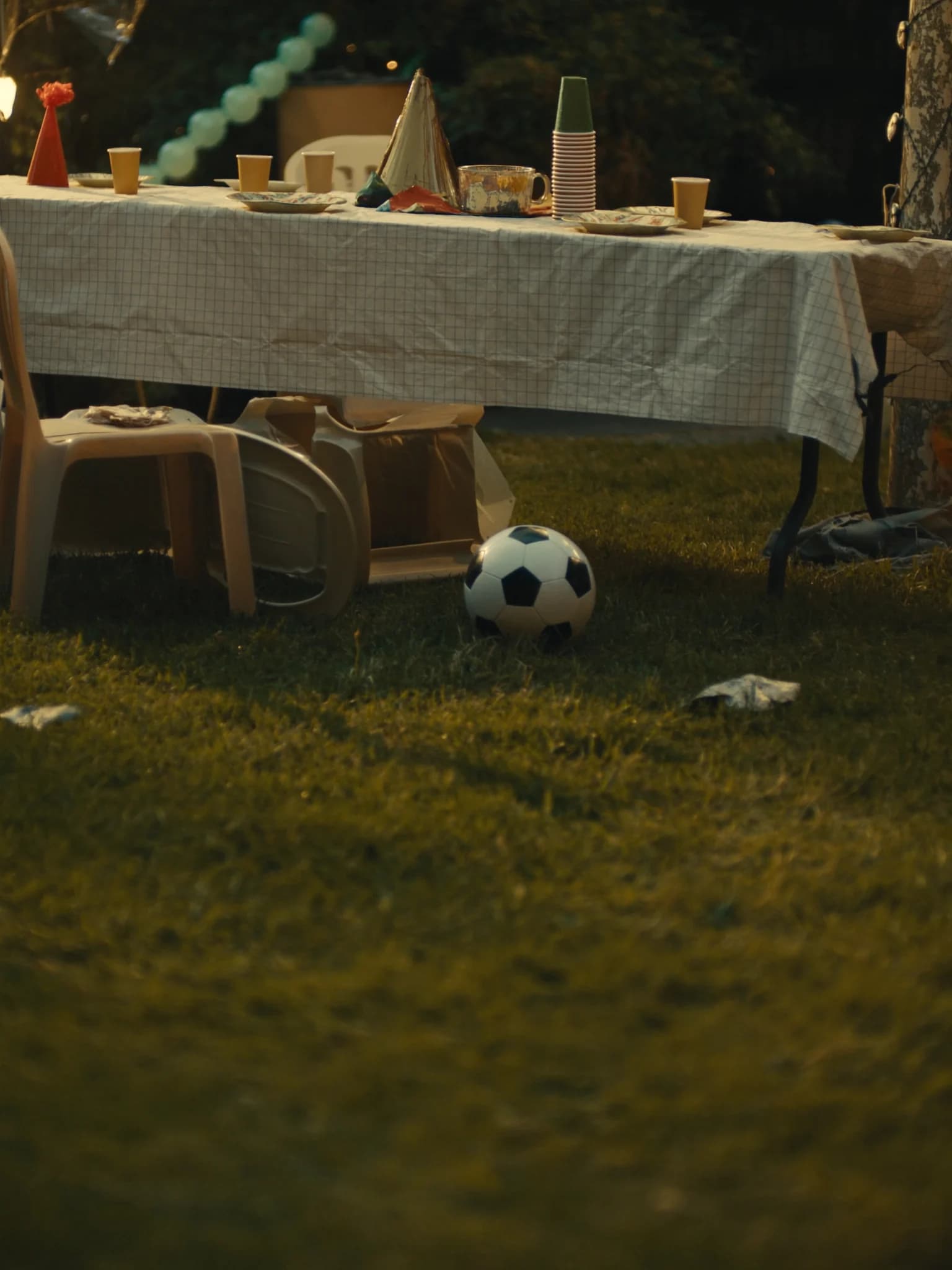 A backyard party table at dusk after the guests have gone, a football left on the grass